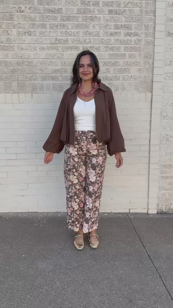 A woman stands outdoors against a white brick wall wearing a brown button-up shirt jacket over a white tank top, floral wide-leg pants, and metallic flat shoes.