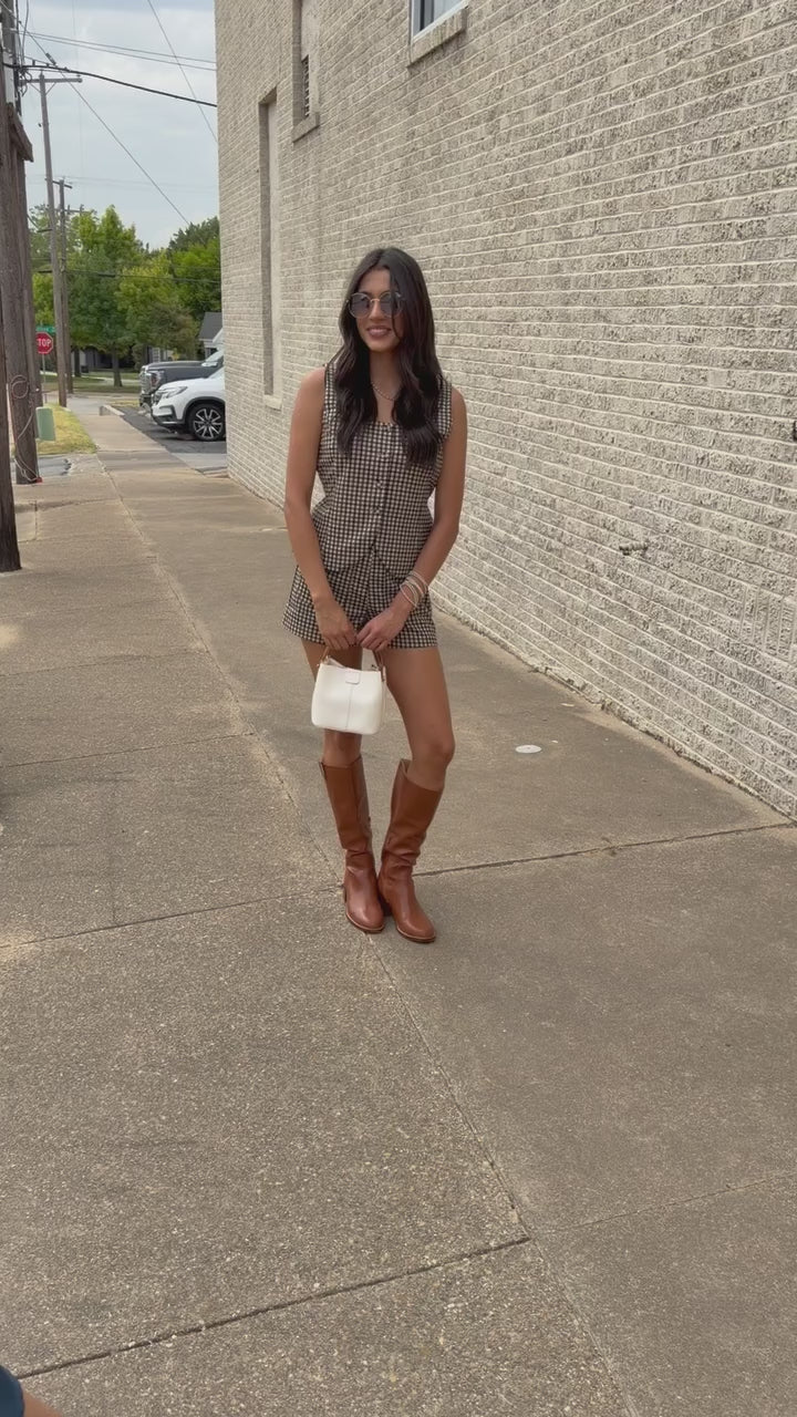 A woman stands outdoors against a light brick wall wearing a sleeveless brown and white gingham romper, brown knee-high boots, and holding a small white handbag.
