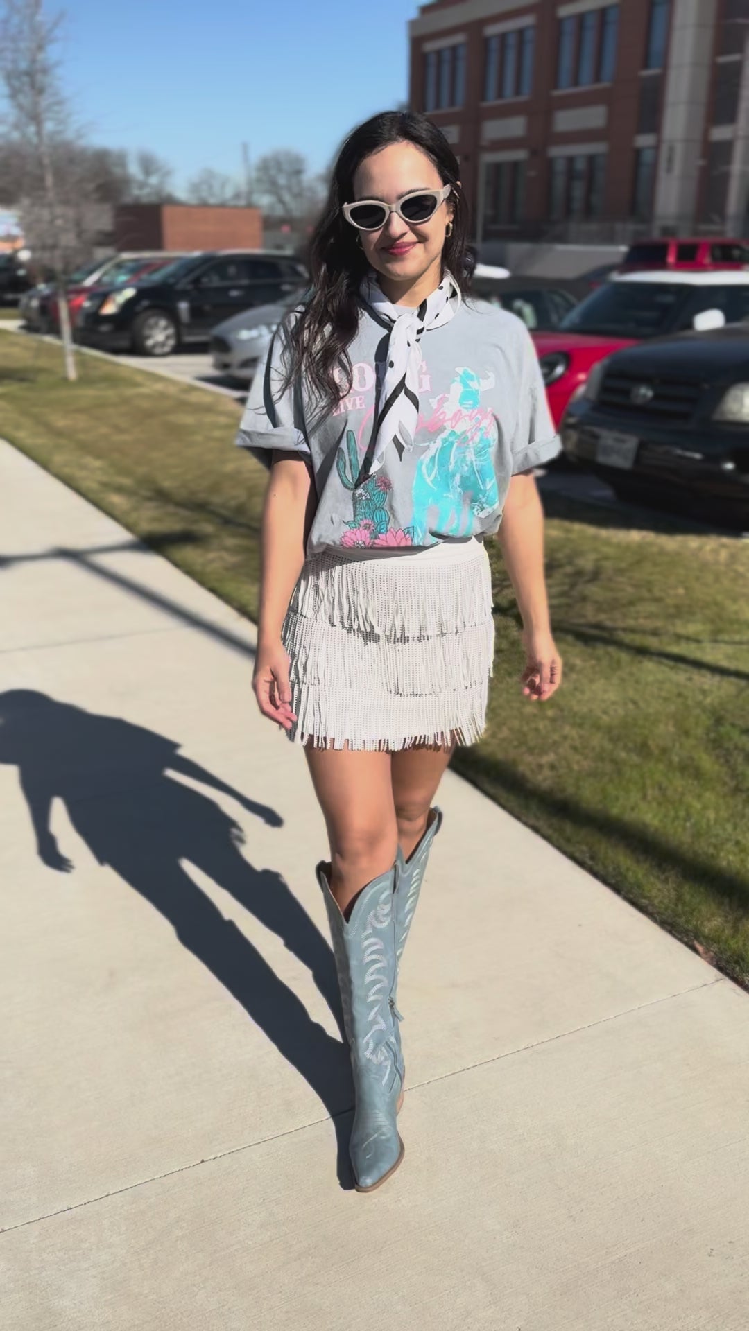 Woman wearing a gray t-shirt with colorful graphics and a white fringe skirt, standing outdoors.