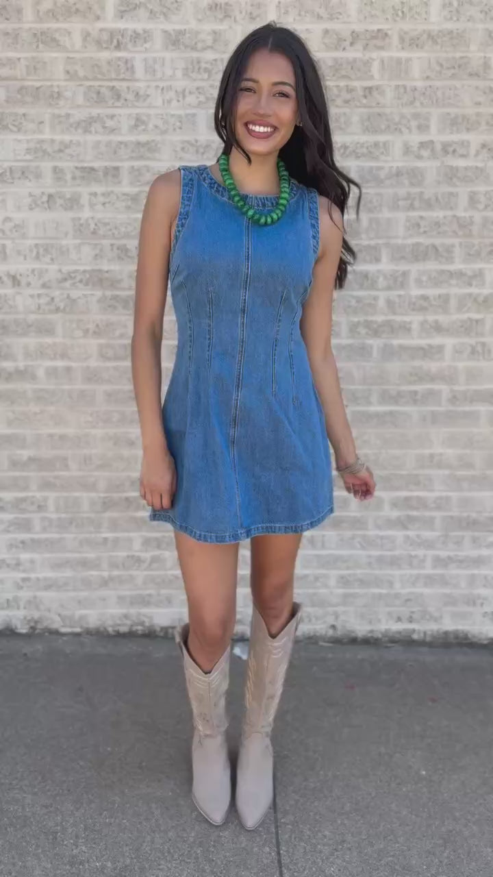 A woman stands on a sidewalk wearing a sleeveless light blue denim mini dress, a chunky green statement necklace, and beige knee-high boots, posing next to a brick wall.
