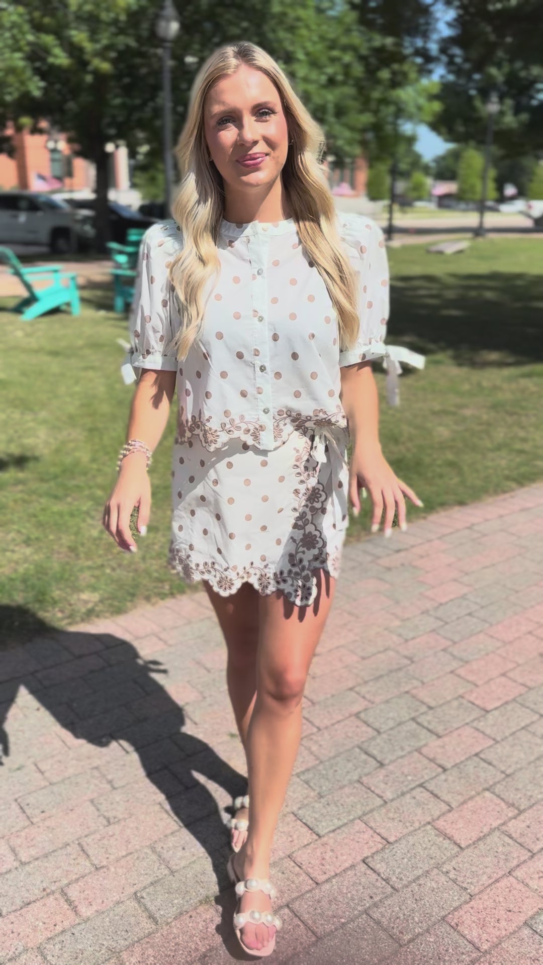 Blonde model standing outside holding a brown purse wearing a cream top and wrap skort set with taupe dots. The set also includes a scalloped hem.