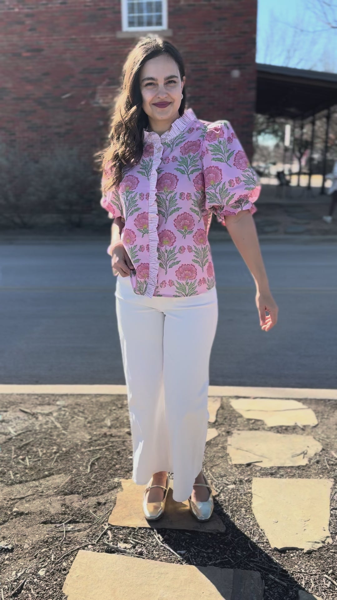 Woman wearing a pink floral blouse and white pants outdoors.