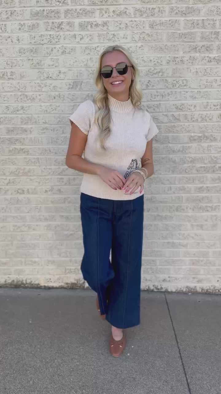 A woman with blonde hair wearing a cream knit sweater with a blue horse graphic and dark blue jeans, standing in front of a white brick wall.