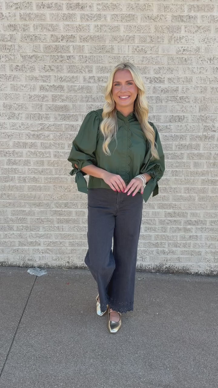 A woman stands on a sidewalk next to a light brick wall, wearing an olive green ruffled blouse with puffed sleeves and dark charcoal wide-leg pants.