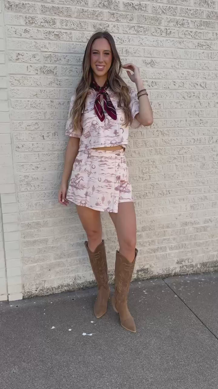A woman stands outdoors on a sidewalk wearing a matching western-style short-sleeve top and skirt set in a light beige print with a dark red neck scarf and tan cowboy boots.