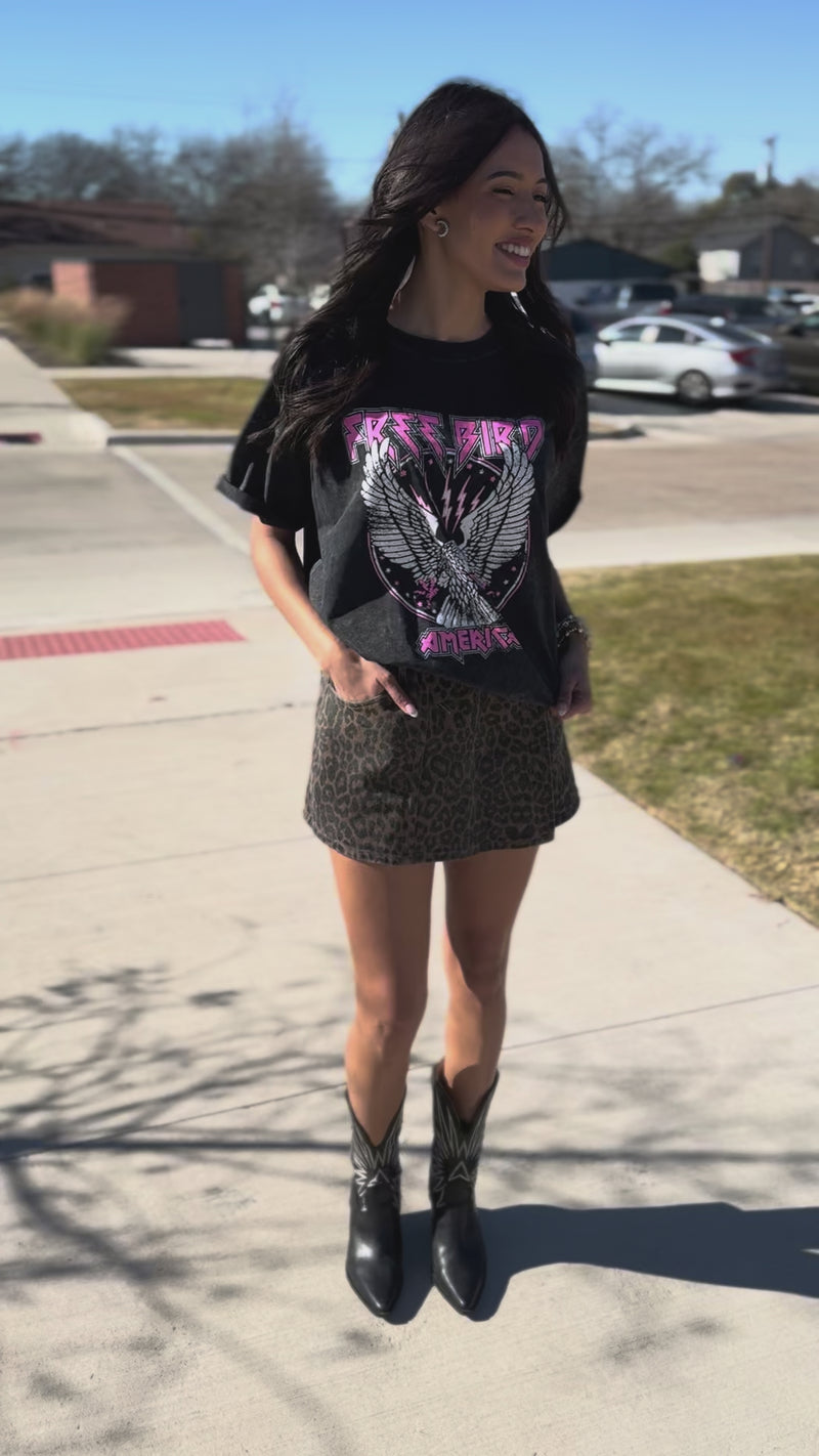 Woman wearing a graphic t-shirt, leopard print skirt, and black boots on a sidewalk.
