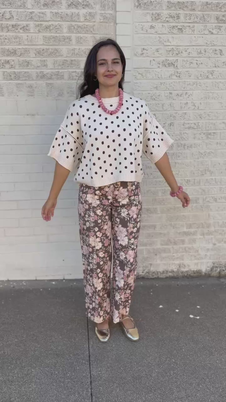 A woman wearing a cream knit sweater with brown polka dots and floral pants, accessorized with a chunky pink beaded necklace and bracelet, standing outdoors next to a white brick wall.