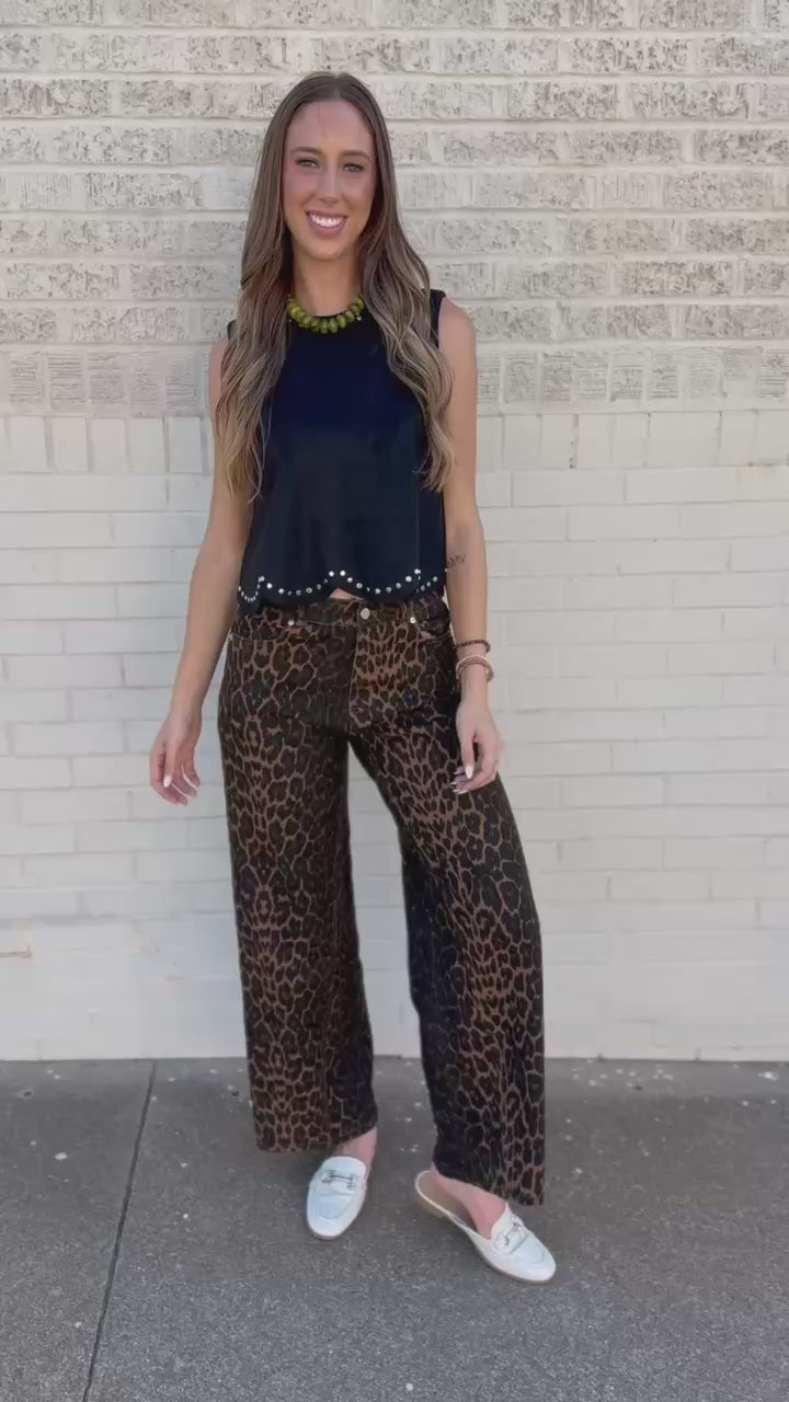 A woman wearing a sleeveless black top with a scalloped hem and white stud embellishments, paired with leopard print pants, standing against a light brick wall.