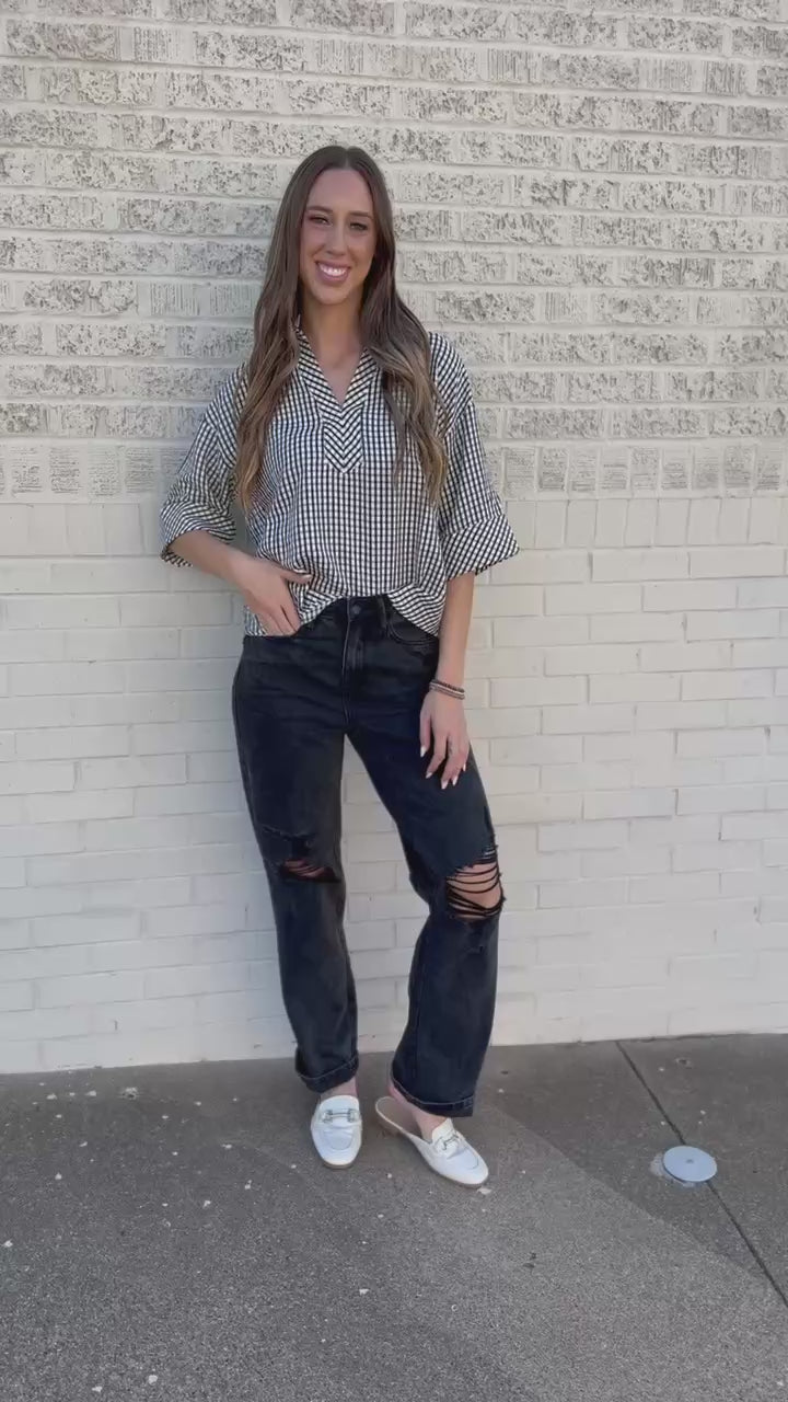 A woman wearing a black and white gingham check short-sleeve blouse and dark denim jeans, standing outdoors in front of a building.