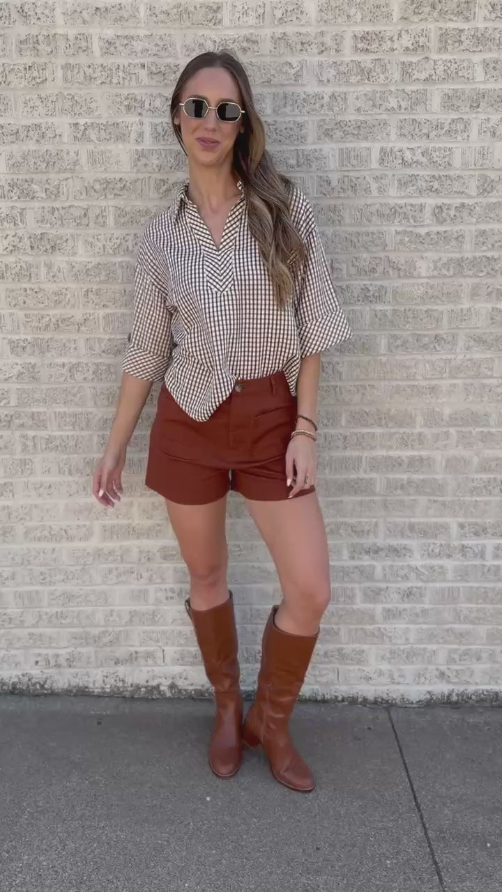 A woman stands outdoors against a light brick wall wearing a beige and white gingham button-up shirt, rust-brown shorts, and brown knee-high boots.