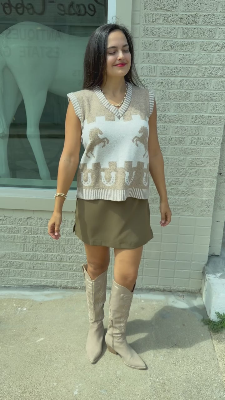 Brunette model, outside, wearing a light mocha sweater vest, tan boots, and a chocolate colored skort.