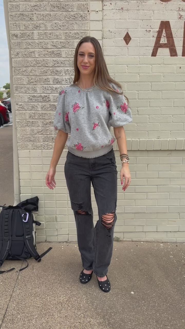 Blonde model wearing a heather grey sweater with rose patterns, black denim pants, and black ballet flats, standing outside. Southern House Clothing