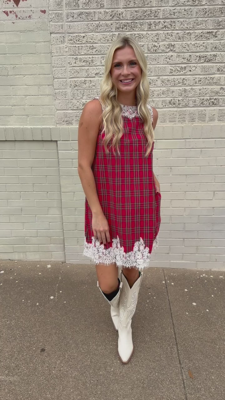 Blonde model standing outside wearing the Hollybrook Plaid Lace Mini Dress in red, green, and black tartan plaid featuring an ivory floral lace neckline and lace-trimmed hem with the white Senora boots from Southern House Clothing