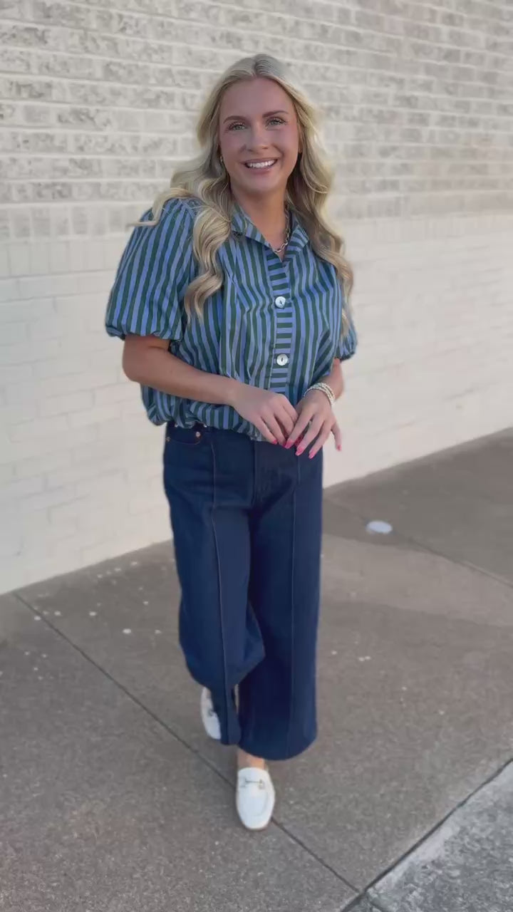 A woman stands outdoors wearing a short-sleeve button-front blouse with vertical teal and white stripes, dark wide-leg jeans, and white slip-on shoes.