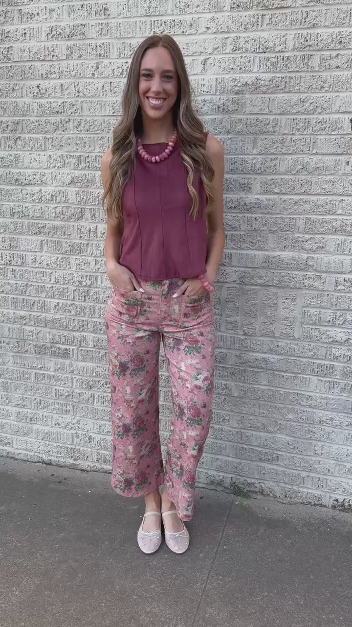 A woman wearing a sleeveless dusty rose top and floral pants, accessorized with a chunky pink beaded necklace and a brown shoulder bag, photographed outdoors.