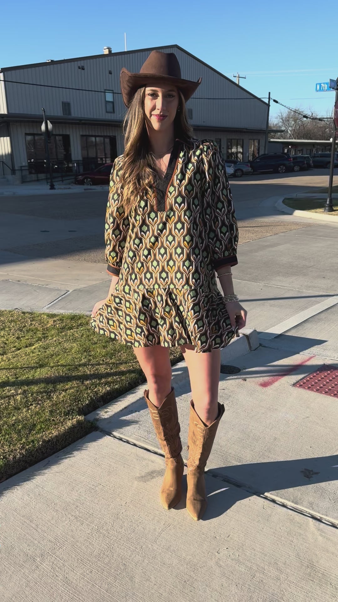 Woman in a patterned dress and cowboy hat standing on a sidewalk.