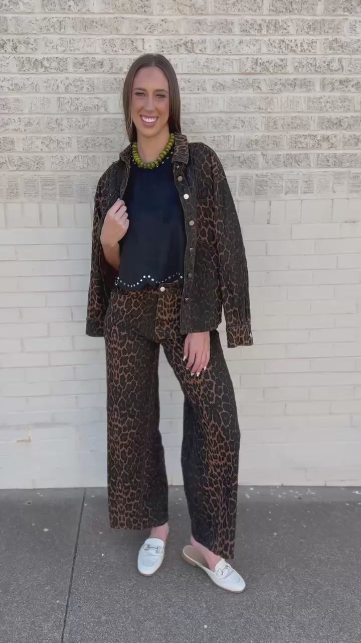 A woman stands against a light beige brick wall wearing a matching leopard-print jacket and wide-leg pants set with a black top and white slip-on shoes.