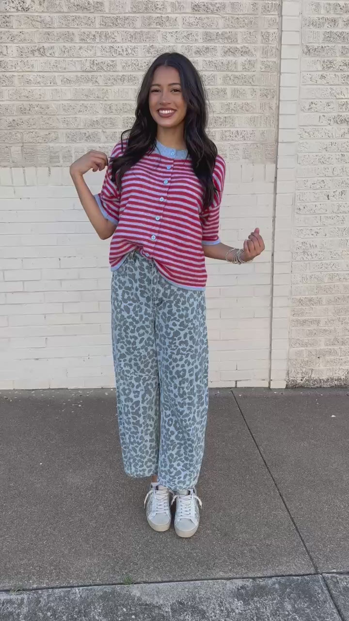 A woman stands outdoors against a light beige brick wall wearing a red and white striped short-sleeve top with blue trim, light blue leopard-print pants, and white sneakers.