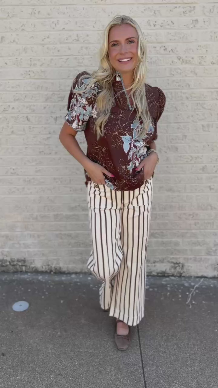 A woman stands on a sidewalk next to a light brick wall, wearing a brown short-sleeve blouse with a white floral print and cream-and-brown vertically striped wide-leg pants.