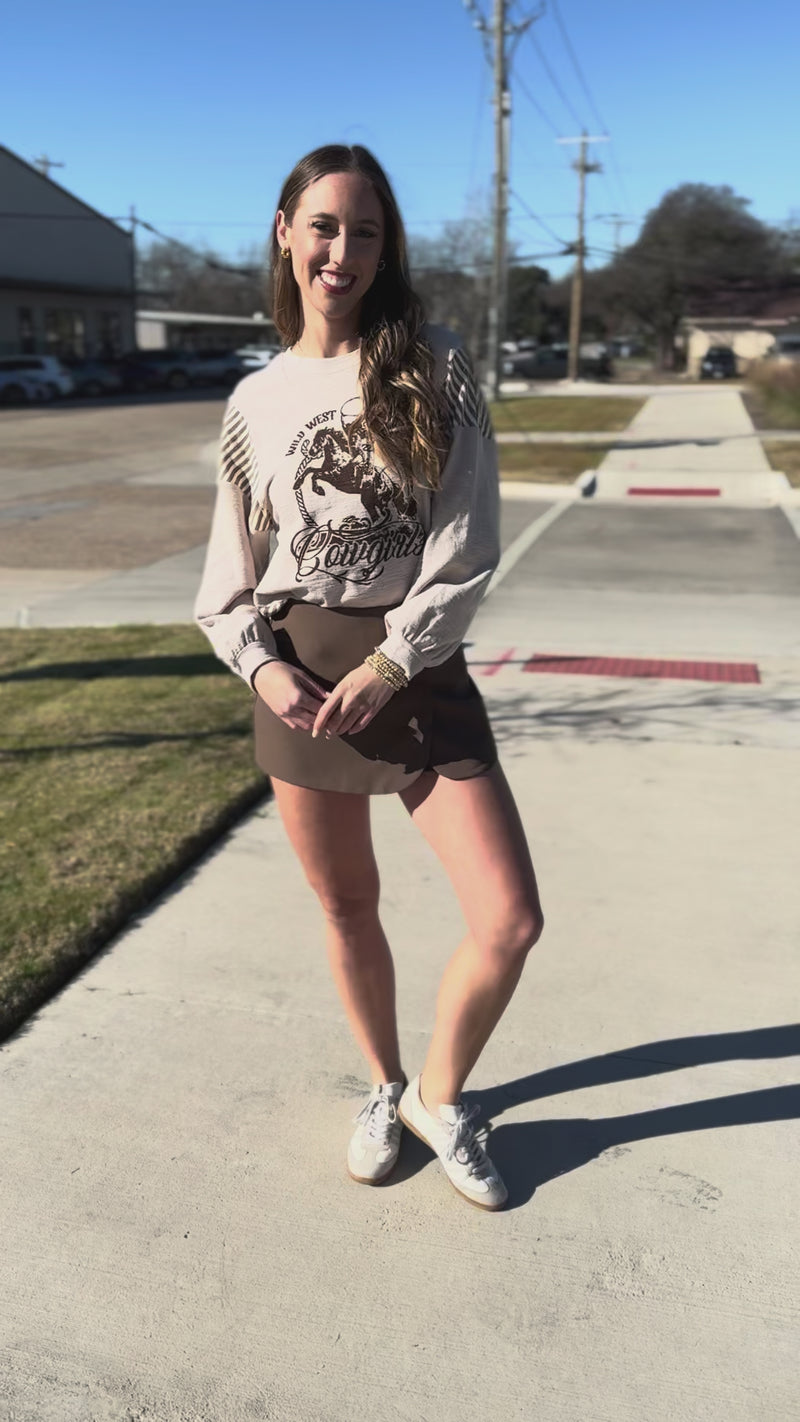 Woman wearing a graphic sweatshirt and brown skirt standing outside.