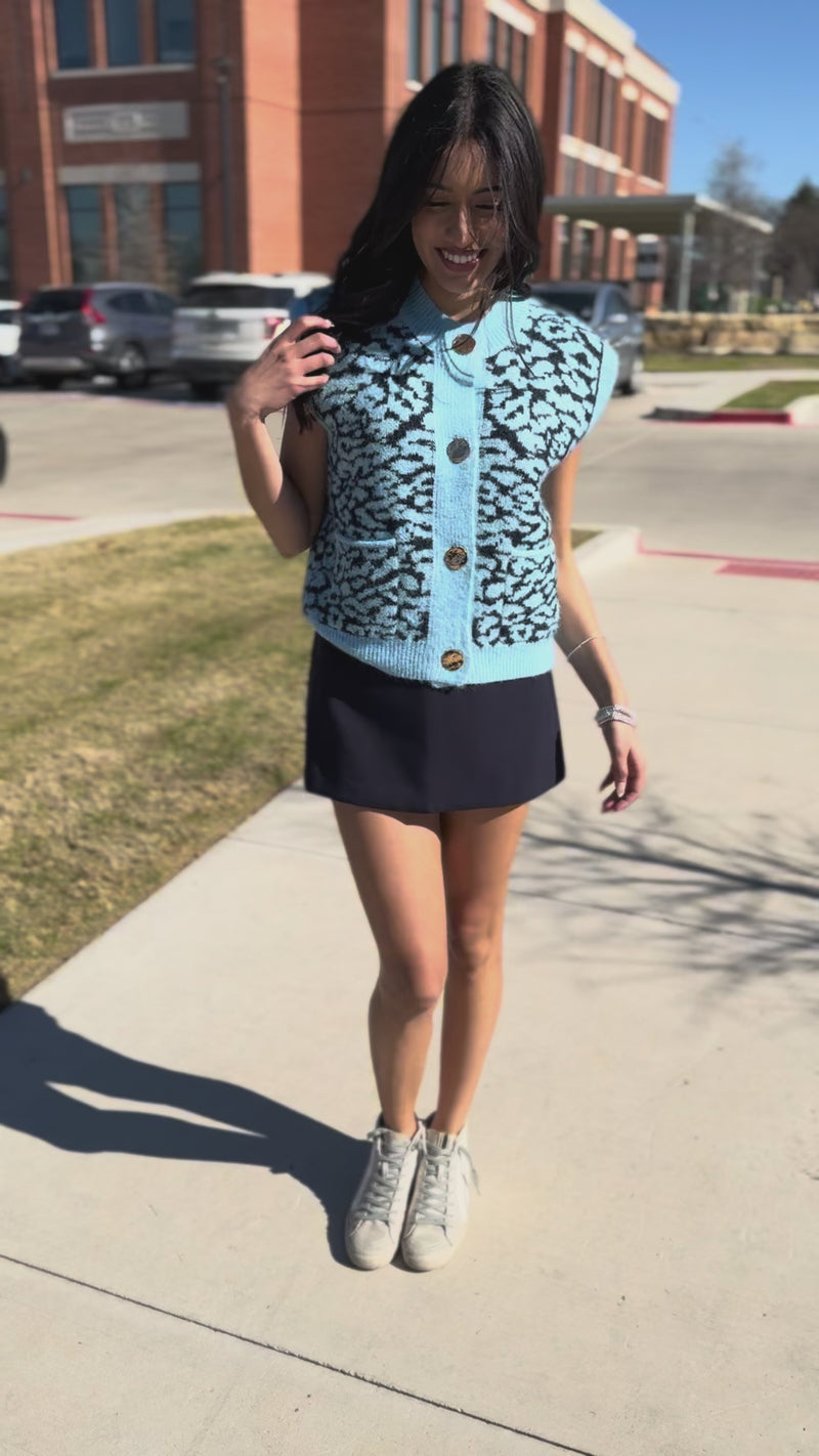 Woman wearing a blue patterned sleeveless top and navy skirt on a city street.