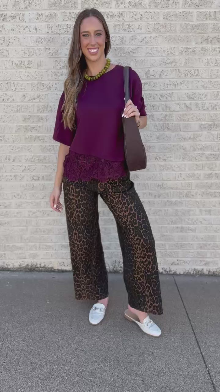 A woman wearing a loose-fitting deep burgundy top and leopard-print pants, carrying a brown shoulder bag, standing outdoors against a light brick wall.