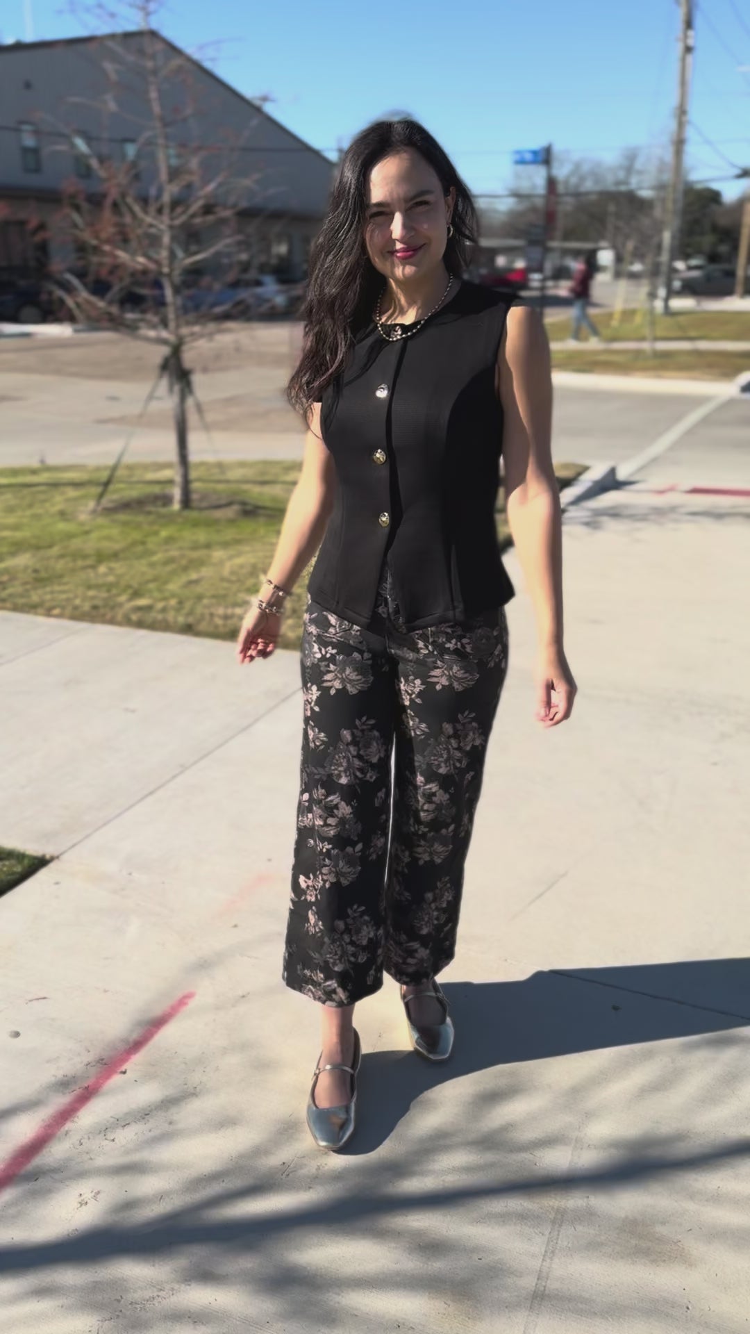 Woman wearing a black sleeveless top with buttons and floral pants outside.