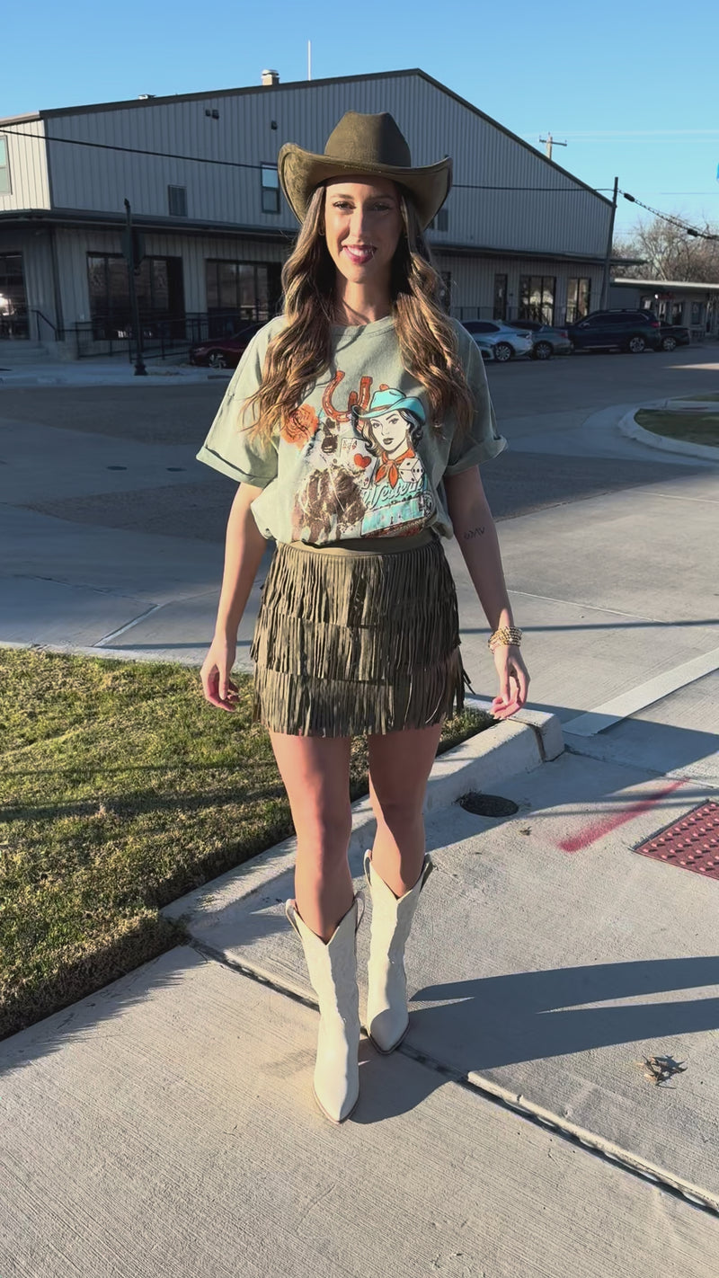 Woman wearing a graphic t-shirt, fringed skirt, and cowboy hat in an outdoor setting.