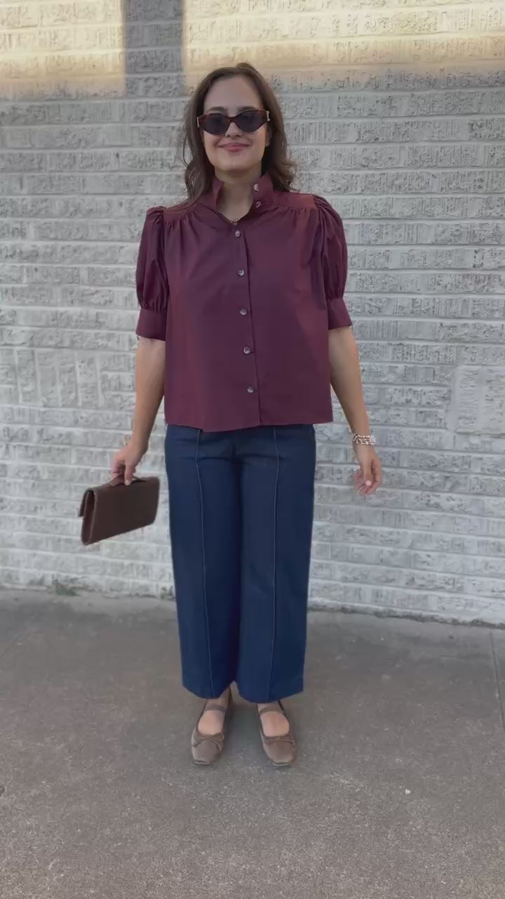 A woman walking outdoors on a sidewalk wearing a burgundy short-sleeve button-up blouse with puffed shoulders, dark wide-leg jeans, brown flats, sunglasses, and holding a brown clutch.
