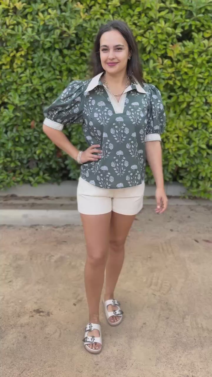 Woman wearing a green floral blouse and white shorts standing outdoors.