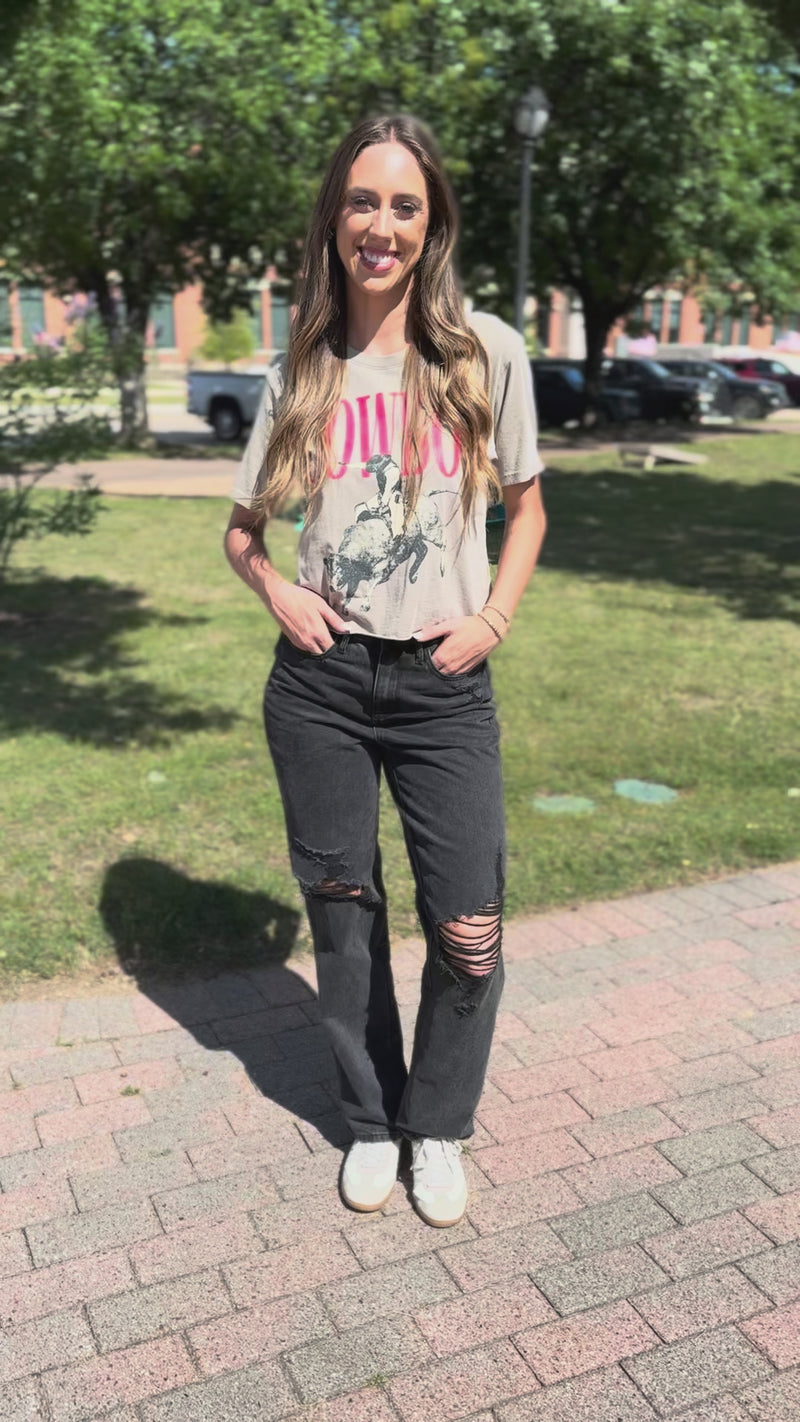 A woman stands on a sidewalk wearing a beige graphic t-shirt with a cowboy and rider print, dark gray distressed jeans, and white sneakers. 