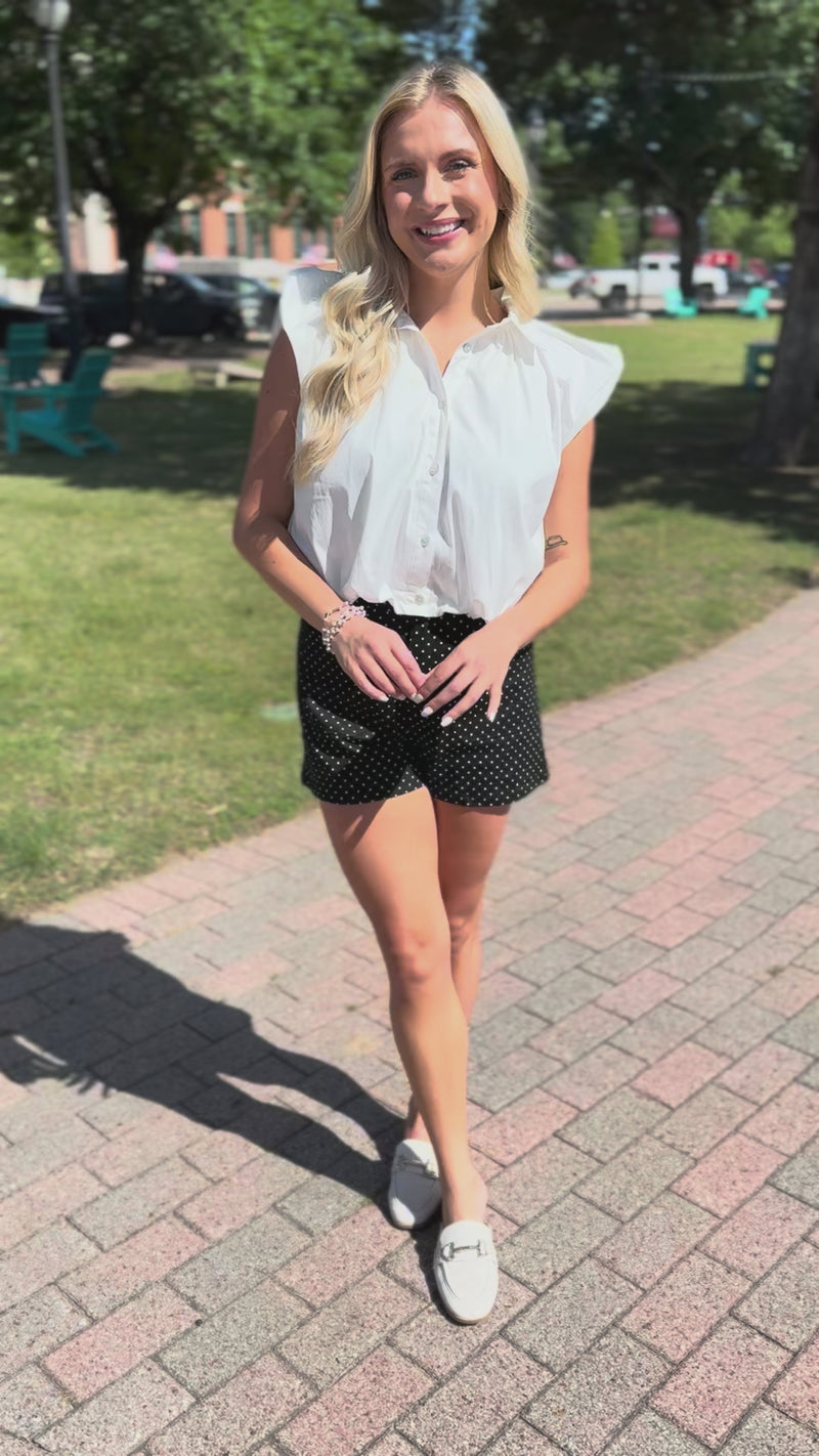Blonde model wearing a sleeveless white top, black shorts with white dots standing outside.