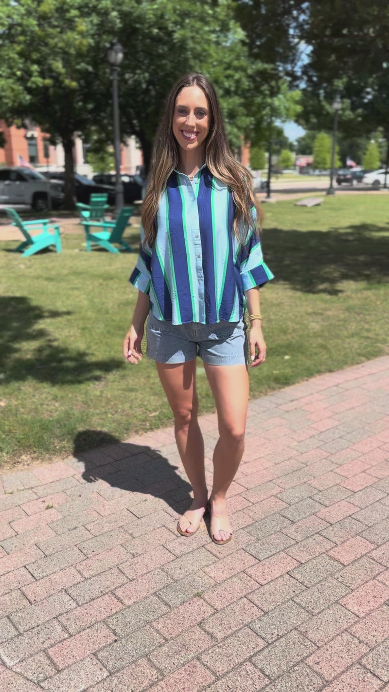 A woman wearing a blue, green, and white striped button-down shirt and denim shorts stands outdoors.