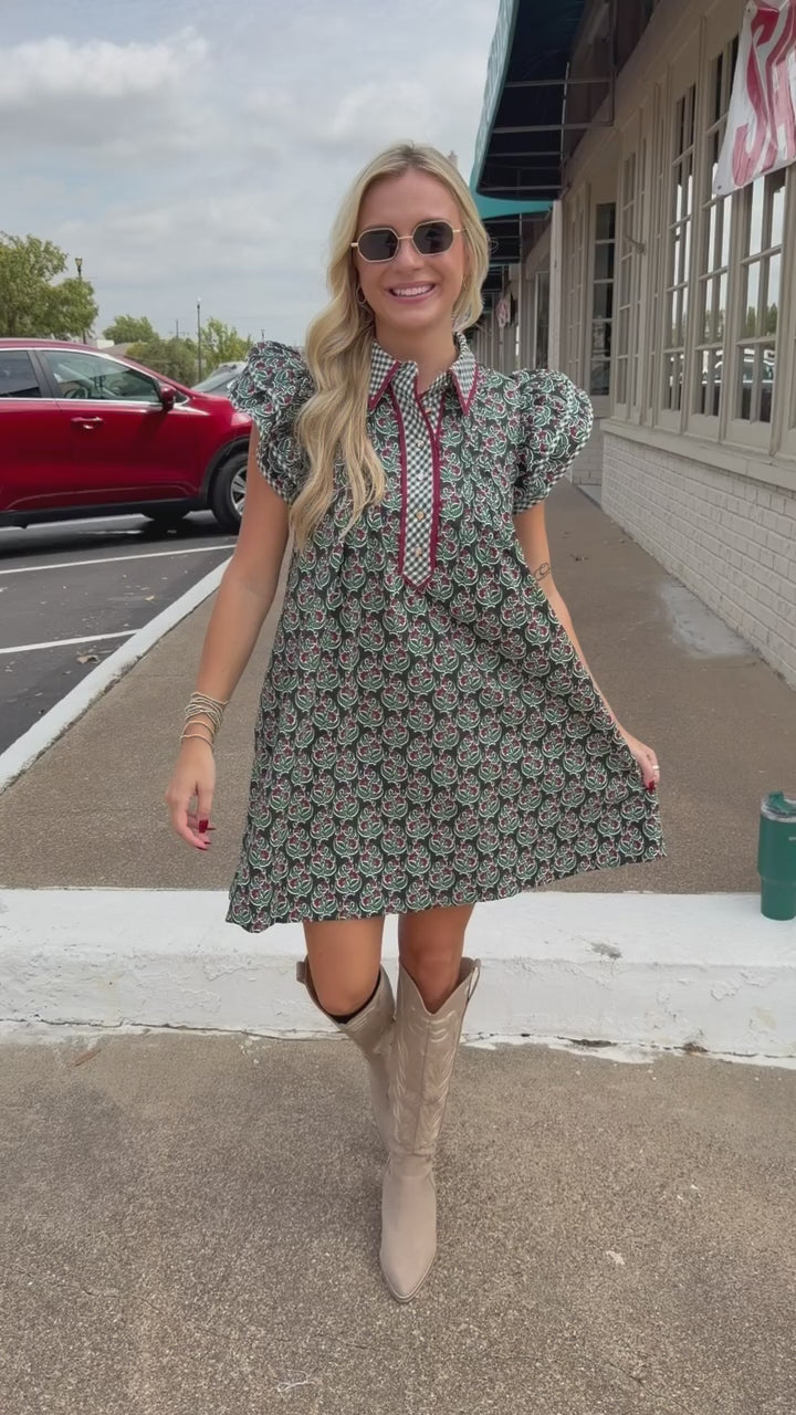 Blonde model standing outside wearing a green dress with red trim and brown cowboys boots. Southern House Clothing