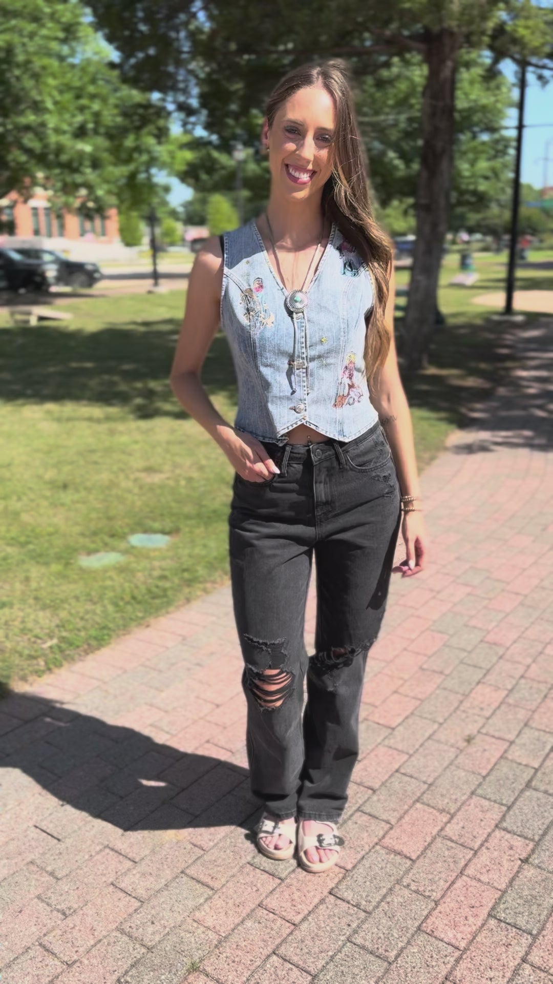 A woman wearing a sleeveless light-wash denim vest with a floral print and distressed black jeans with ripped knees, walking on a sidewalk.