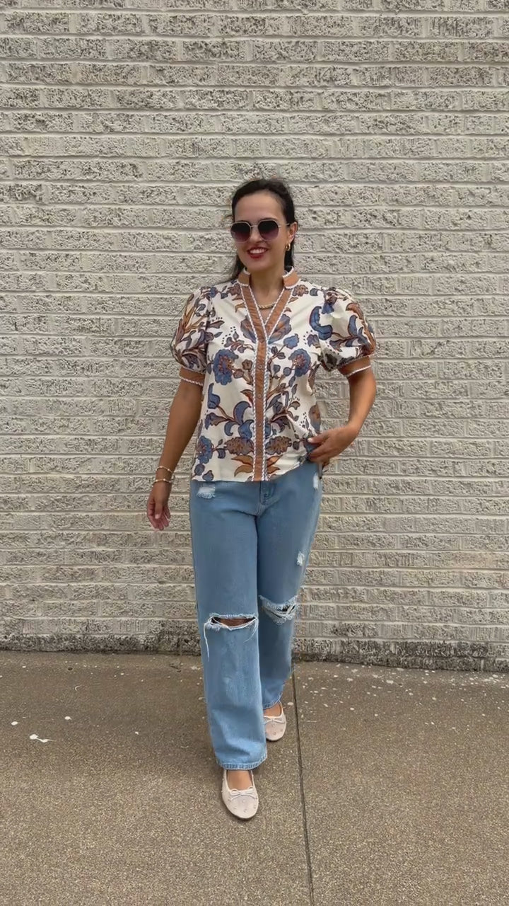 Brunette model wearing a ecru, tan, and blue floral blouse with sunglasses on and denim pants, standing outside. Southern House Clothing