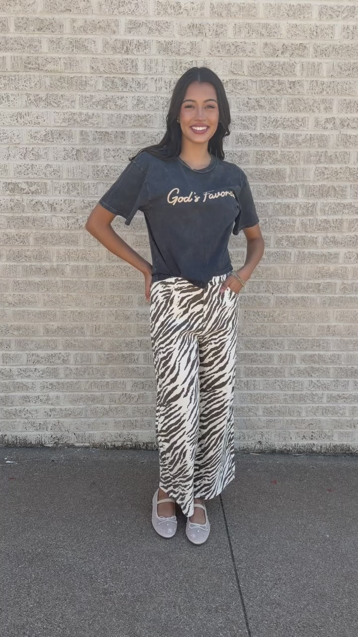 A woman standing on a sidewalk wearing a dark gray graphic t-shirt with 'God's Favorites' printed on the chest and high-waisted, wide-leg zebra-print pants in brown and white.