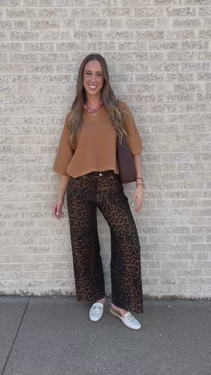 A woman stands on a sidewalk against a light brick wall, wearing a tan short-sleeve blouse, leopard-print pants, and carrying a dark brown crescent shoulder bag.