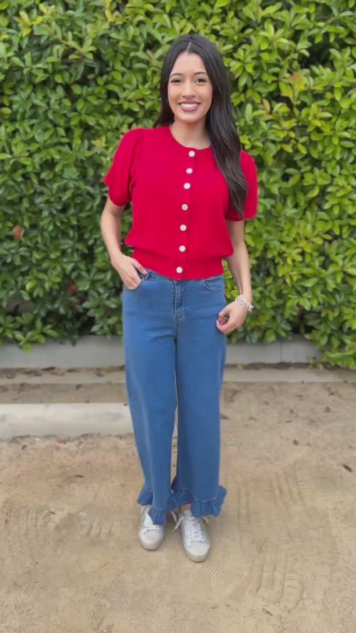 Woman wearing a red blouse with white buttons and blue jeans, standing outdoors.