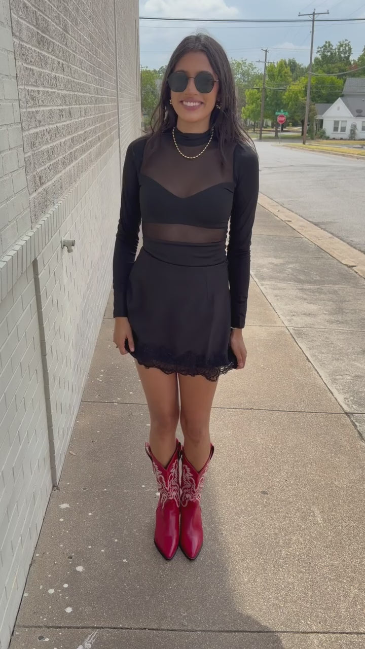 Model wearing Nocturne Mesh Panel Bodysuit in Black featuring long sleeves and sheer mesh panels, styled with Bellarose Lace Hem Mini Skort in Black and red western boots standing outside. Southern House Clothing