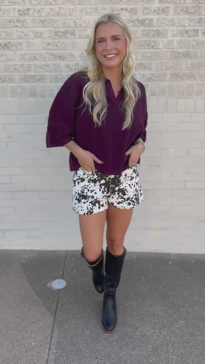 A woman modeling a loose-fitting deep purple short-sleeve top and black-and-white patterned shorts with black knee-high boots, standing against a light beige brick wall.
