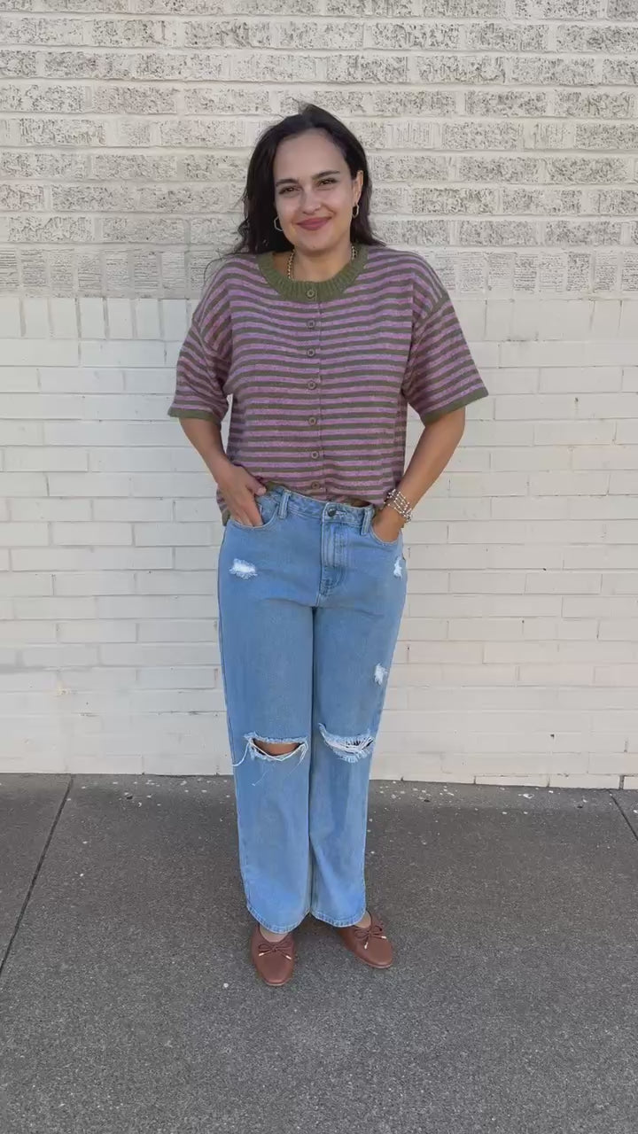 A woman wearing a short-sleeve knit henley top with horizontal olive green and dusty rose stripes, paired with light-wash blue jeans, standing outdoors on a sidewalk.