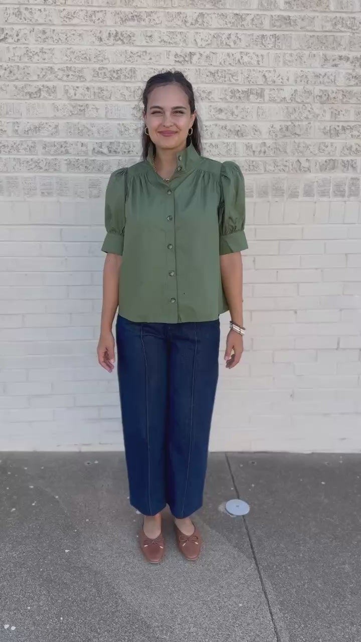 A woman stands on a city sidewalk wearing a sage green short-sleeve button-up blouse with puff sleeves and dark navy wide-leg pants.