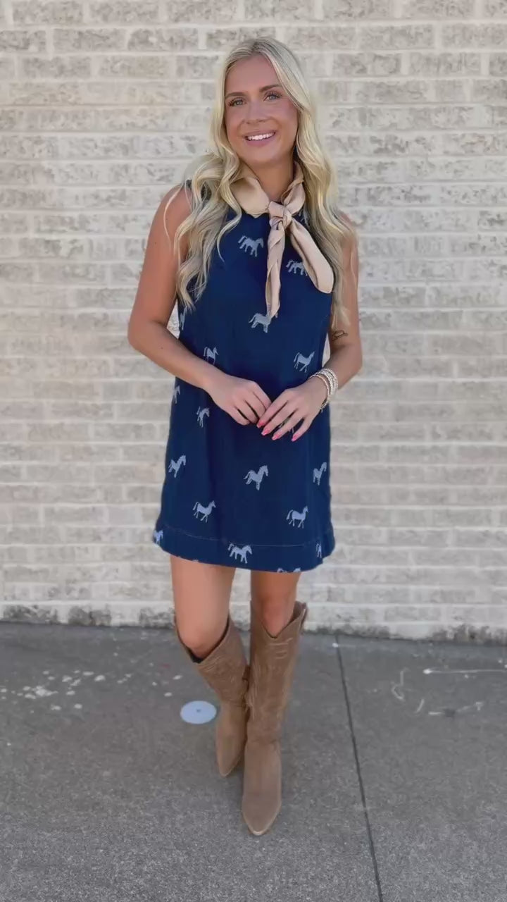 Blonde model standing outside wearing a sleeveless, denim dress with horse prints and brown cowboy boots.