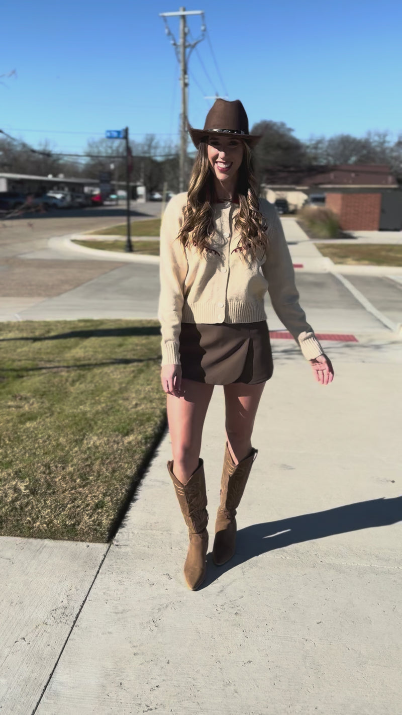 Woman wearing a beige cardigan with red accents, brown shorts, and a cowboy hat outside.