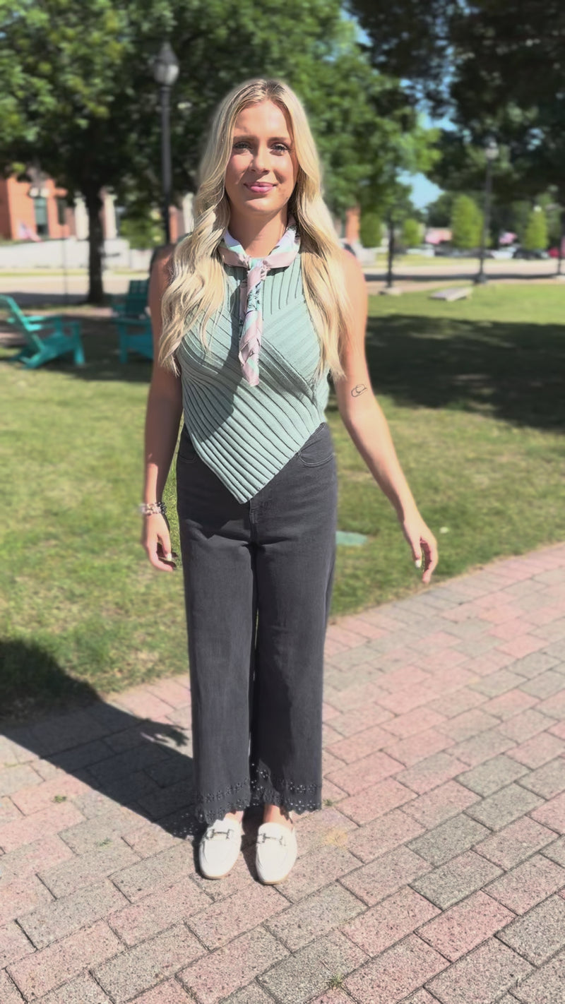 A smiling blonde woman wearing a sleeveless ribbed mint green top with a light floral neck scarf, paired with dark gray jeans, standing outdoors