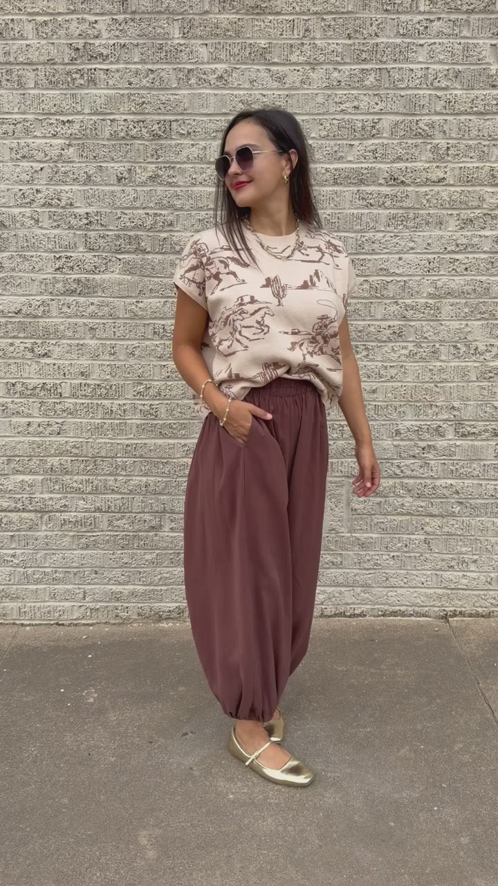 Model wearing Bronco Trail Knit Top in Ecru featuring taupe western cowboy, bucking horse, and desert cactus illustrations, styled with the Tulara Barrel Pants – Brown from Southern House Clothing.