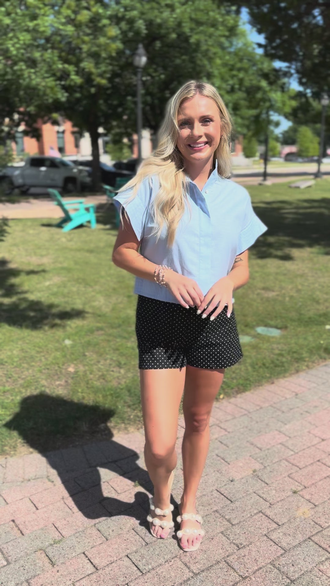 Blonde model wearing a light blue blouse and black shorts with white dots, standing outside.
