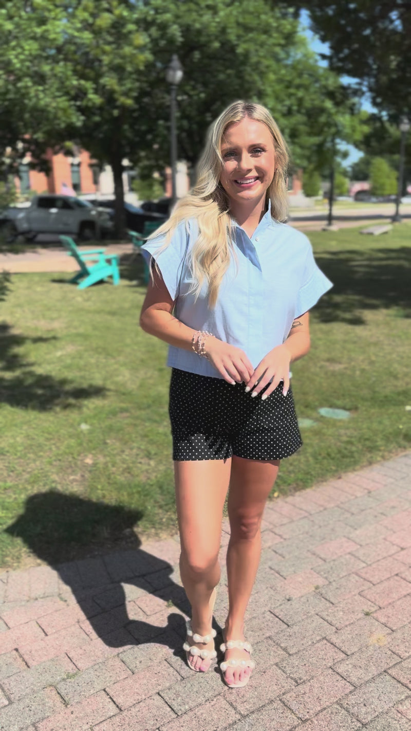 Blonde model wearing a light blue blouse and black shorts with white dots, standing outside.