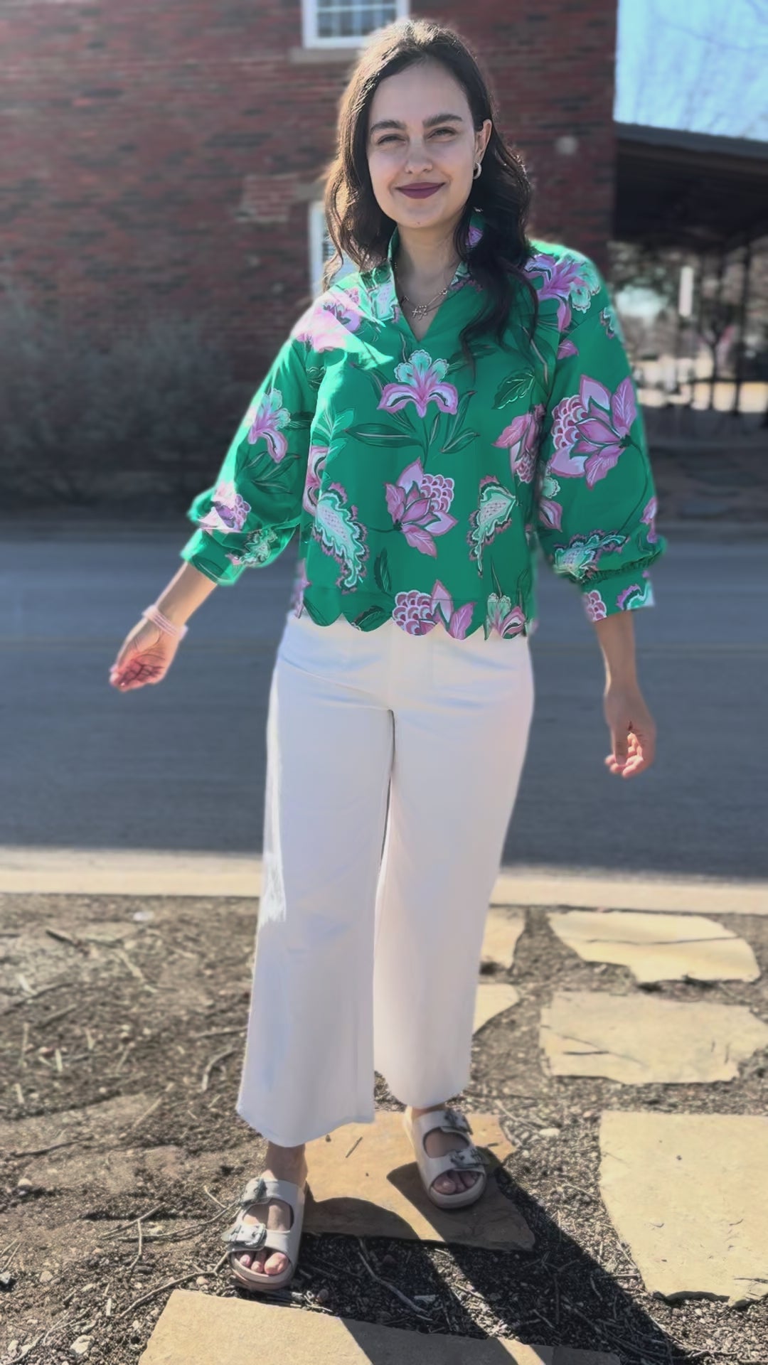 Woman wearing a green floral blouse and white pants on a sidewalk.